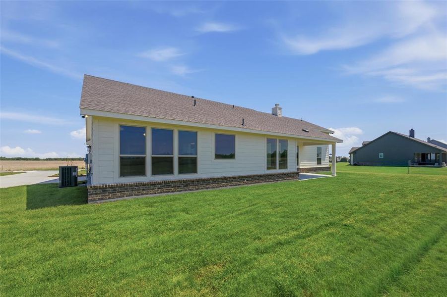 Rear exterior featuring a covered patio, extensive green lawn, and a light-colored siding facade with a dark stone-look skirting Rear exterior featuring a covered patio, extensive green lawn, and a light-colored siding facade with a dark stone-look skirting