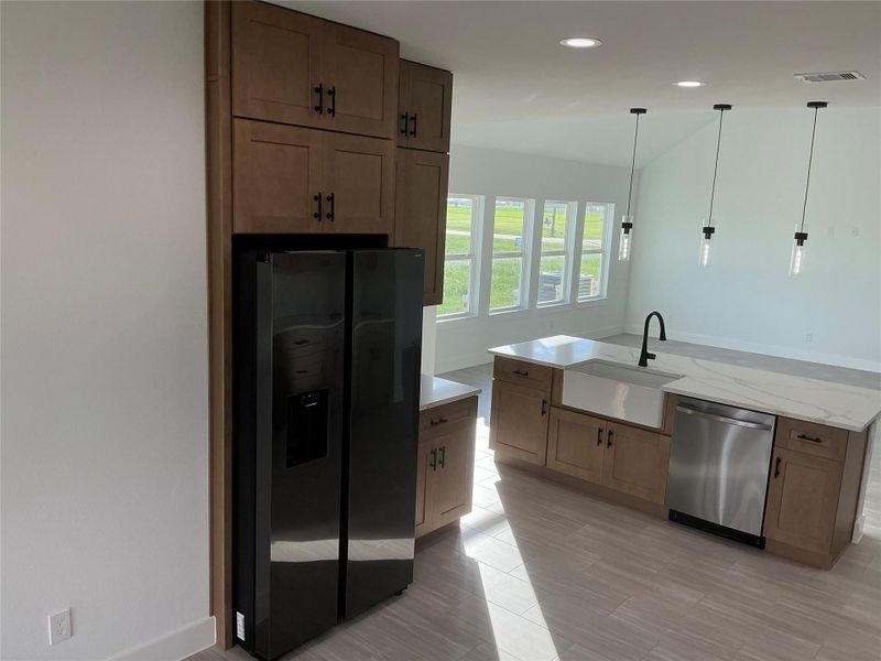 This modern kitchen features sleek black appliances, wooden cabinetry, and a spacious island with a farmhouse sink. The open layout is enhanced by large windows allowing natural light and contemporary pendant lighting.