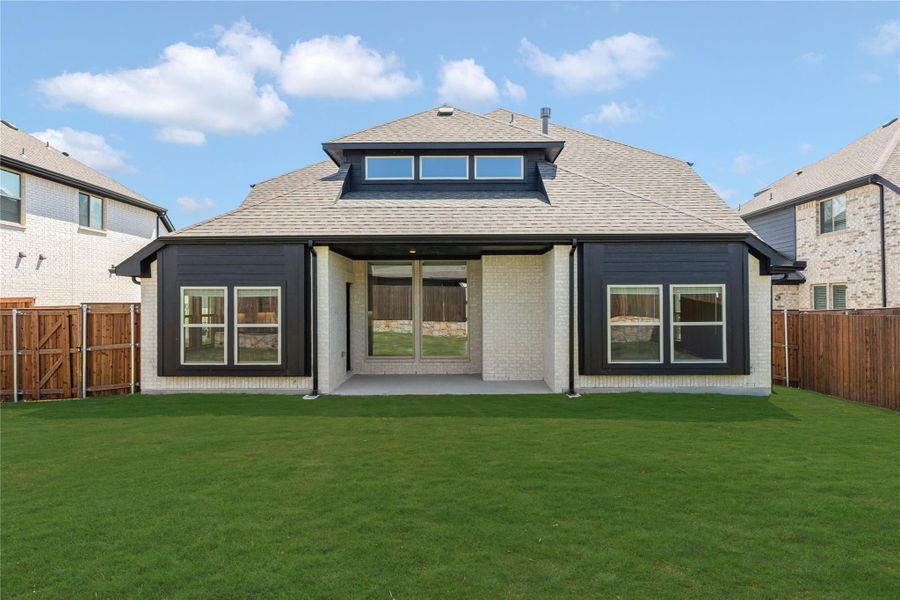 Rear exterior of single-story home with white brick, dark siding accents, covered patio, and large fenced backyard with green lawn.