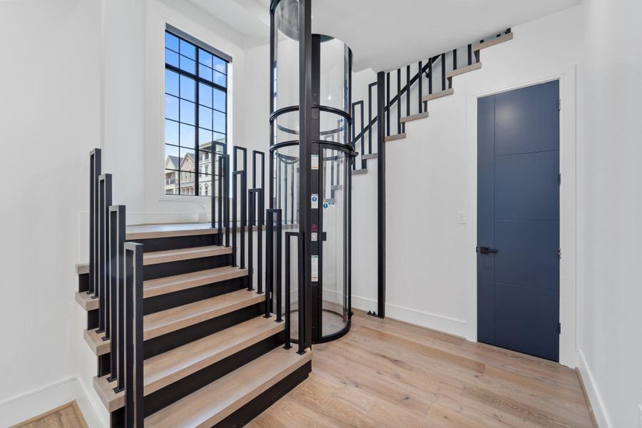 This modern entryway showcases a sleek staircase with black metal railings and light wood treads, elegantly paired with a cylindrical glass elevator. A large picture window floods the space with natural light, while a bold blue door adds a refined touch of contrast. This modern entryway showcases a sleek staircase with black metal railings and light wood treads, elegantly paired with a cylindrical glass elevator. A large picture window floods the space with natural light, while a bold blue door adds a refined touch of contrast.