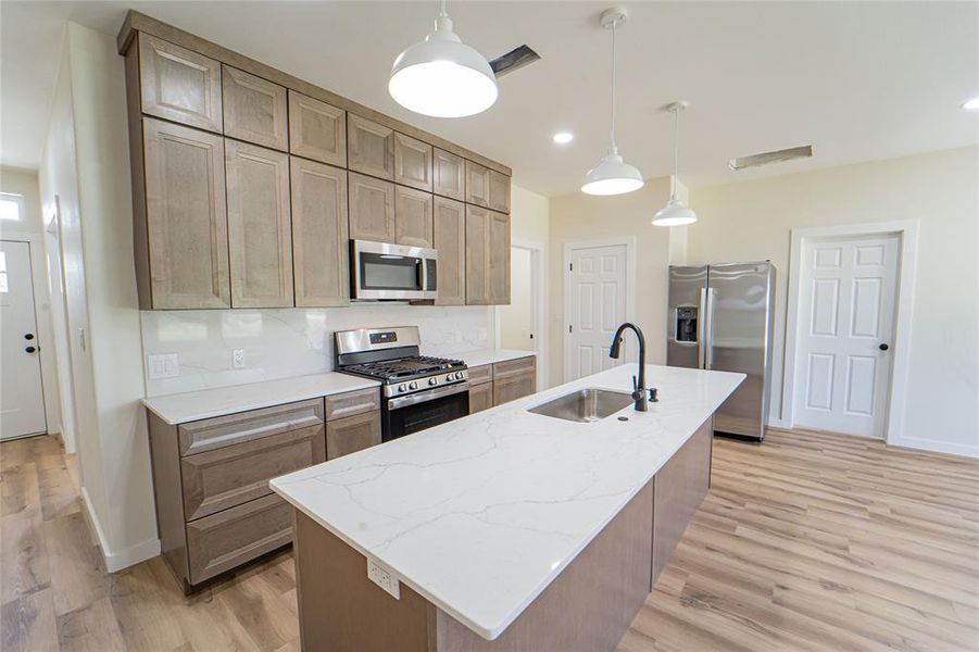 Modern kitchen featuring an expansive center island with an undermount sink, pendant lighting, wood-finish flooring, stainless steel appliances, and shaker-style cabinetry