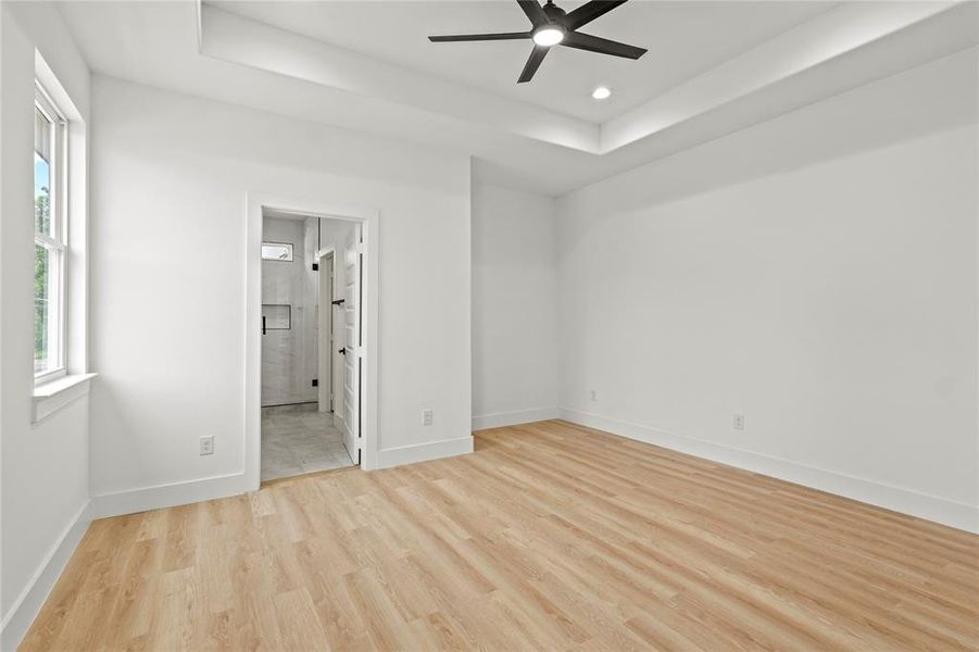 Unfurnished bedroom featuring a raised ceiling, light wood-style flooring, baseboards, recessed lighting, and ensuite bath Unfurnished bedroom featuring a raised ceiling, light wood-style flooring, baseboards, recessed lighting, and ensuite bath
