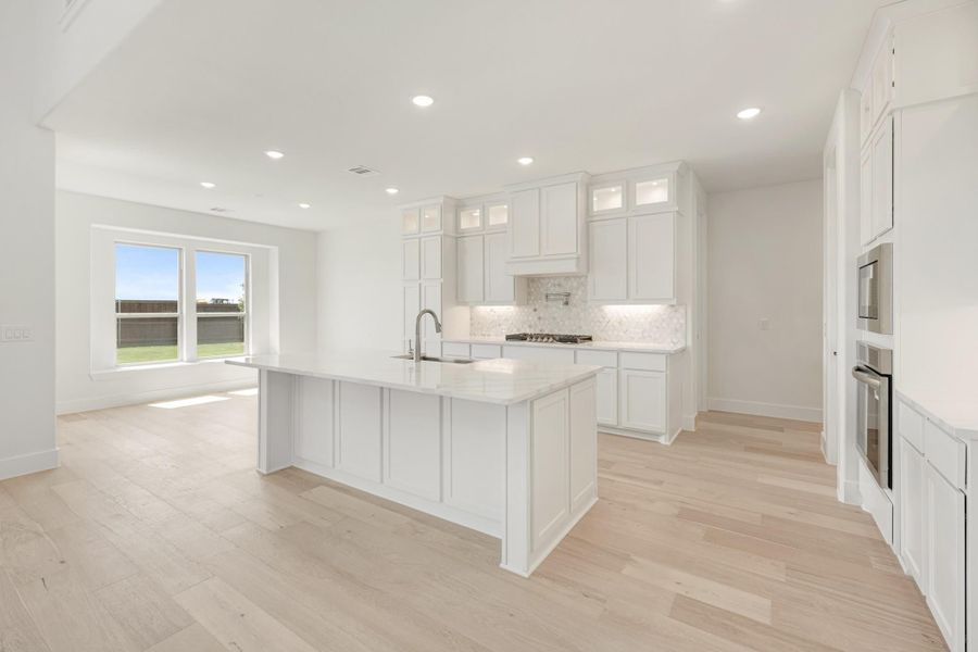 White kitchen with large island, white cabinets, tile backsplash, and light hardwood floors open to living area.