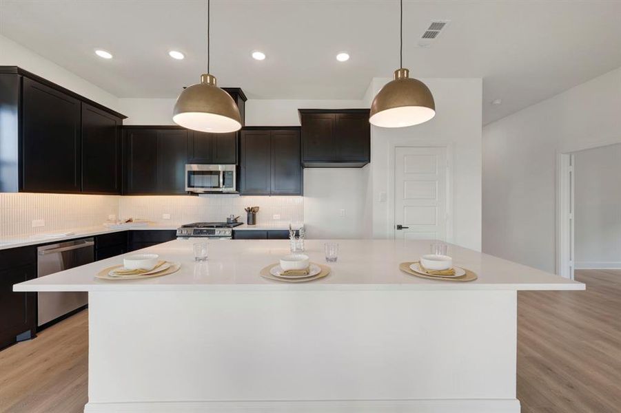 Kitchen with light wood finished floors, a center island with sink, stainless steel appliances, decorative light fixtures, and backsplash Kitchen with light wood finished floors, a center island with sink, stainless steel appliances, decorative light fixtures, and backsplash
