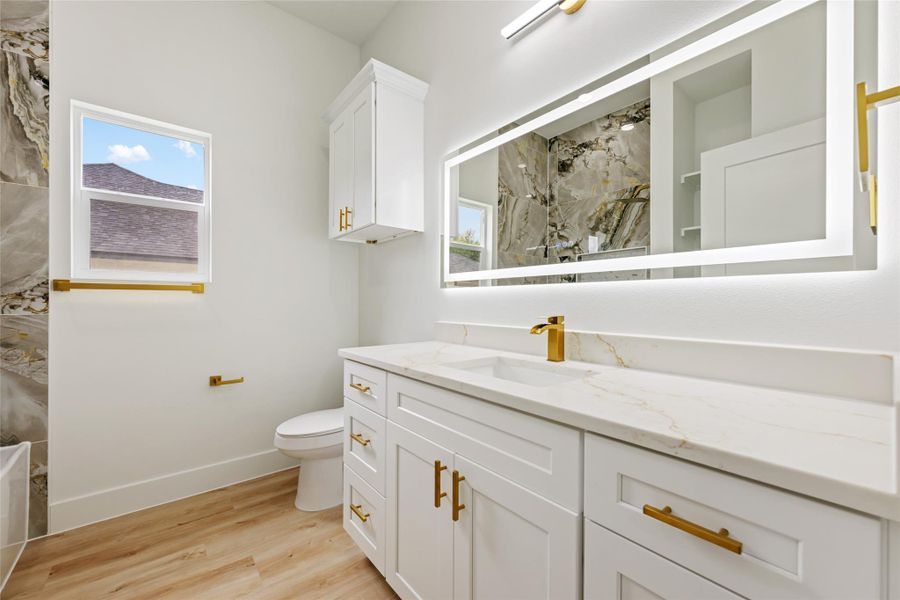 Bathroom featuring a dual-sink vanity with white cabinetry and gold-finish hardware, a built-in illuminated mirror, and wood-finish flooring