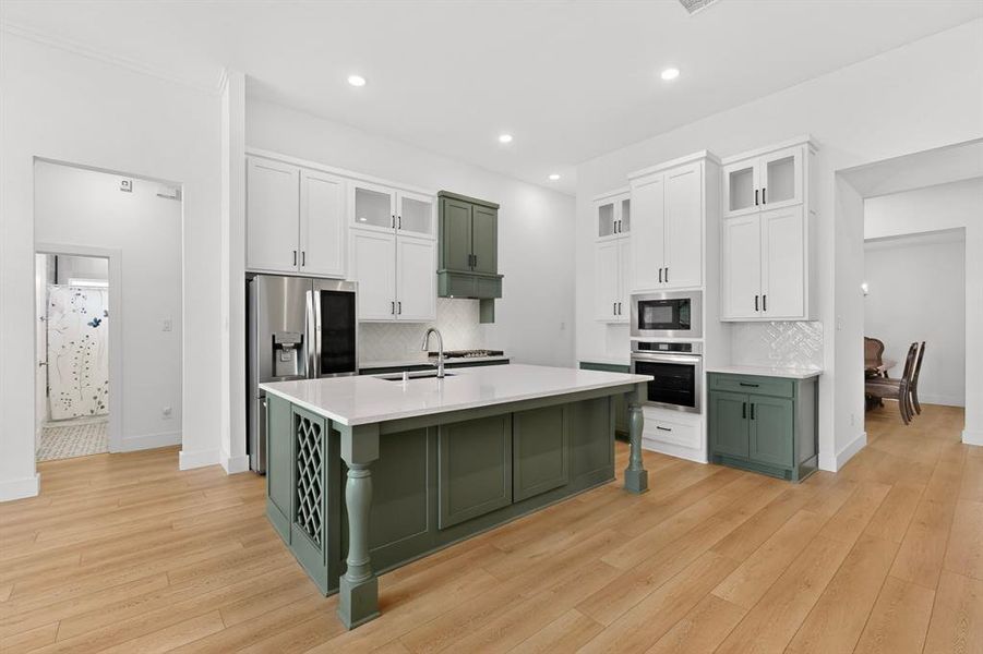 Bright kitchen featuring a central island with white countertop, stainless steel appliances, white cabinetry with glass inserts, green lower cabinetry, and wood-finish flooring Bright kitchen featuring a central island with white countertop, stainless steel appliances, white cabinetry with glass inserts, green lower cabinetry, and wood-finish flooring