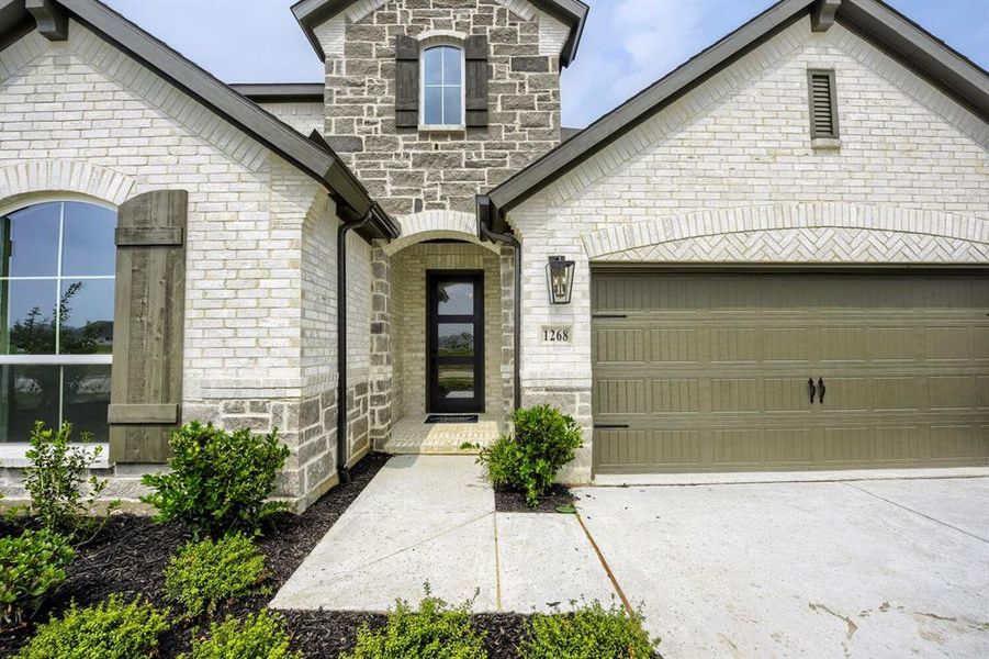 Stone and brick facade featuring a covered entry, arched window openings, dark wood shutters, and an attached garage with a carriage-style door Stone and brick facade featuring a covered entry, arched window openings, dark wood shutters, and an attached garage with a carriage-style door