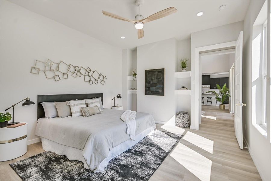 Sun-filled primary bedroom with built-in shelving, high ceilings, and light hardwood flooring.