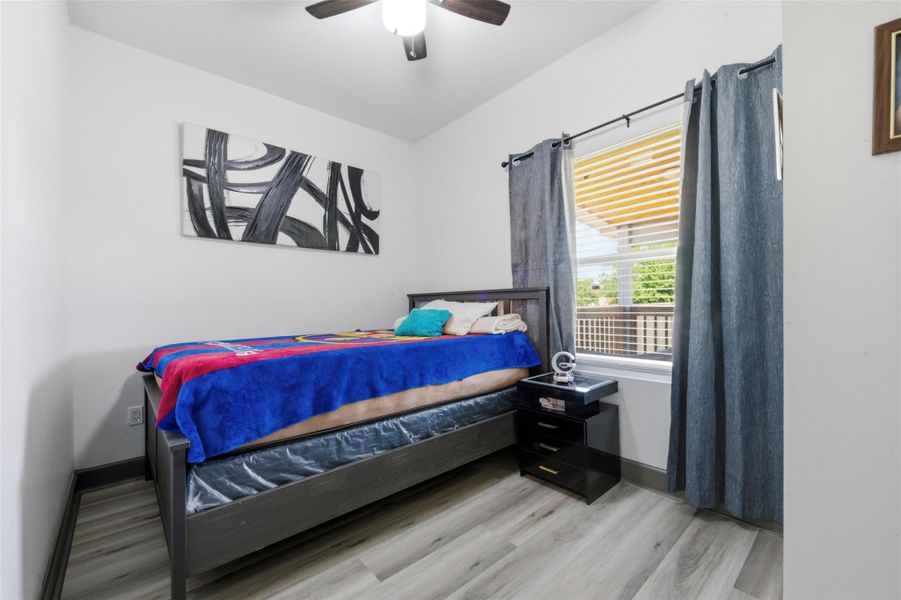 This cozy bedroom features a single bed, and natural light from a window with gray curtains. The room has light wood flooring, creating a welcoming atmosphere.