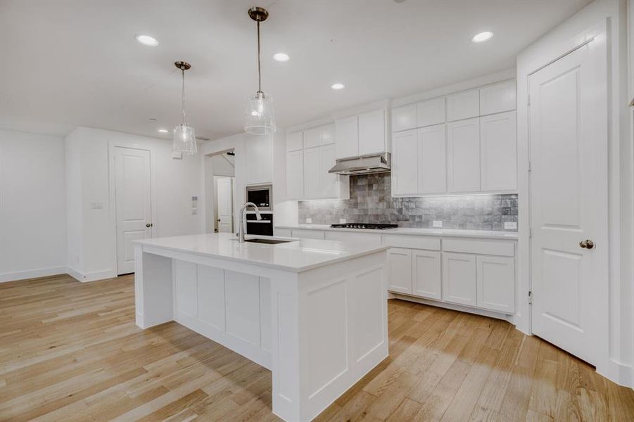 Modern kitchen featuring a large central island with a sink, white cabinetry, integrated stainless steel appliances, a tiled backsplash, and wood-finish flooring