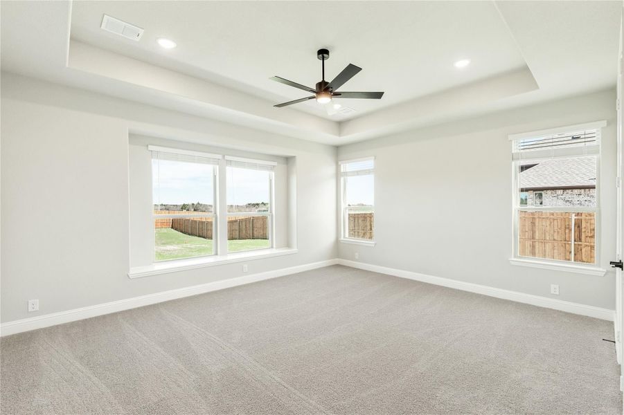 Empty bedroom with tray ceiling, ceiling fan, carpet flooring, and multiple windows overlooking fenced backyard Empty bedroom with tray ceiling, ceiling fan, carpet flooring, and multiple windows overlooking fenced backyard