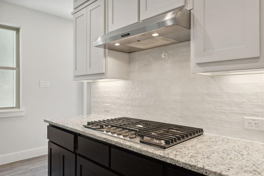 Kitchen featuring under cabinet range hood, baseboards, backsplash, light stone countertops, and stainless steel gas stovetop