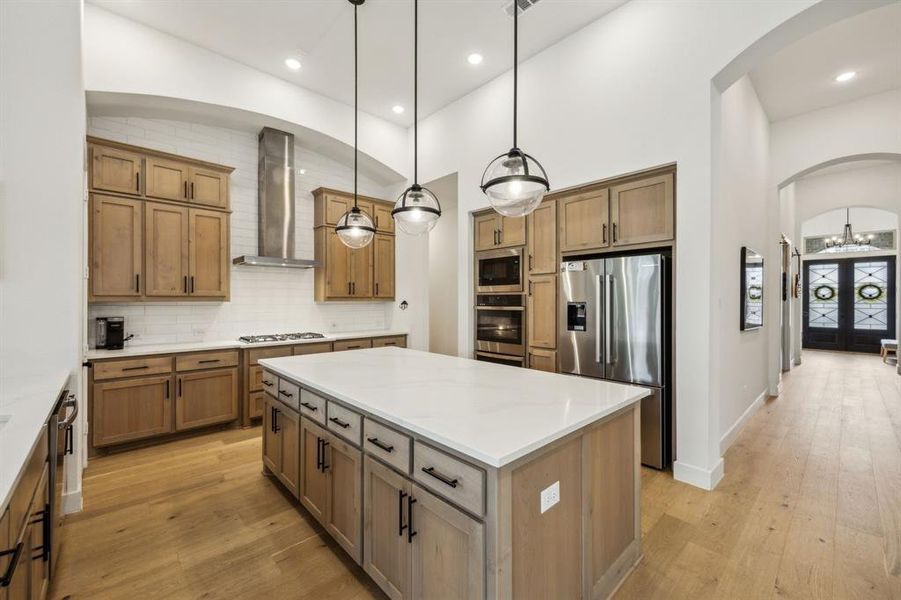 Kitchen featuring stainless steel appliances, wall chimney range hood, arched walkways, a kitchen island, and light wood-style floors