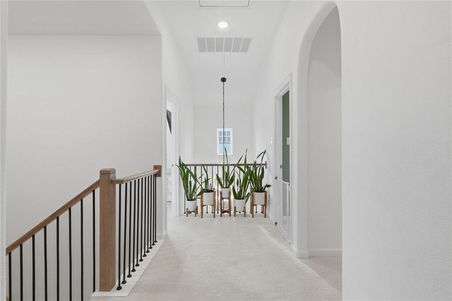 Upper-level hallway featuring light-toned carpet flooring, a stairway with wood-finish handrails and black metal balusters, and a recessed arched doorway Upper-level hallway featuring light-toned carpet flooring, a stairway with wood-finish handrails and black metal balusters, and a recessed arched doorway