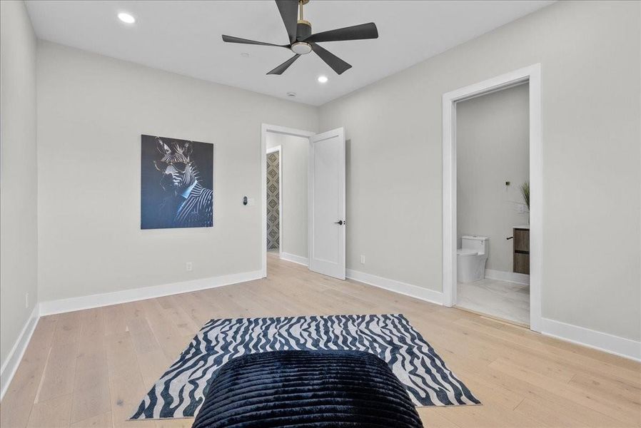 This spacious bedroom features light hardwood floors, a modern ceiling fan, and neutral walls. It includes an ensuite bathroom and a walk-in closet. Artwork and a stylish rug add a touch of personality. This spacious bedroom features light hardwood floors, a modern ceiling fan, and neutral walls. It includes an ensuite bathroom and a walk-in closet. Artwork and a stylish rug add a touch of personality.