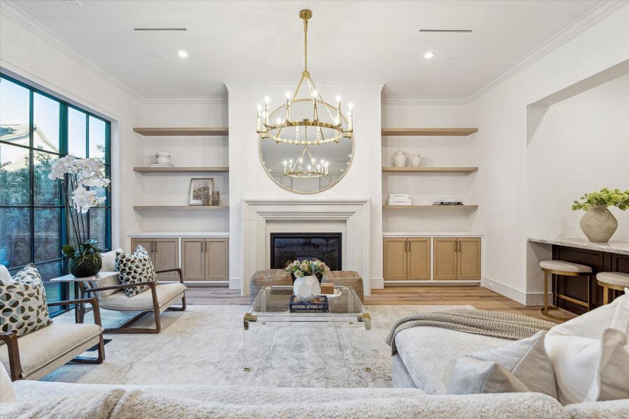 This palatial living room with 11-foot ceilings features a gas log fireplace with a custom cast-stone surround. White oak shelves flank the fireplace for display of art pieces and storage of AV equipment. To the right is a sprawling wet bar, and large format windows flood the space with natural light.