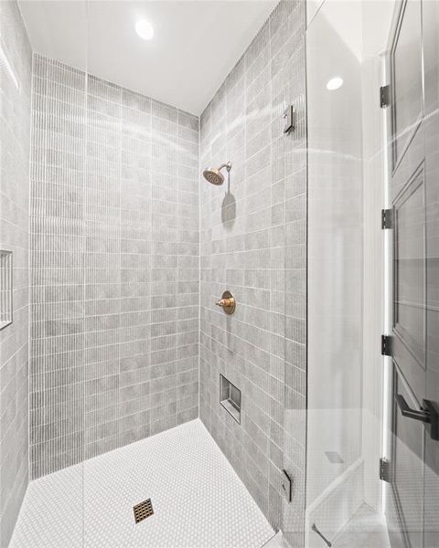 Shower with gray tile walls, mosaic tile floor, recessed niche, brass-finish fixtures, and a clear glass enclosure