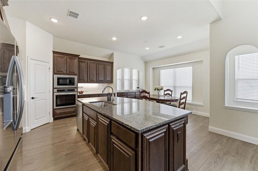 Kitchen featuring dark wood finish cabinets, stainless steel appliances, light stone countertops, light wood-type flooring, and a kitchen island with sink