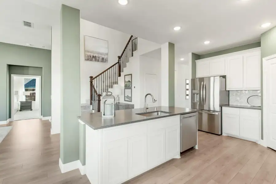 Kitchen with white cabinets, gray countertop island, stainless steel appliances, and light wood floors open to staircase