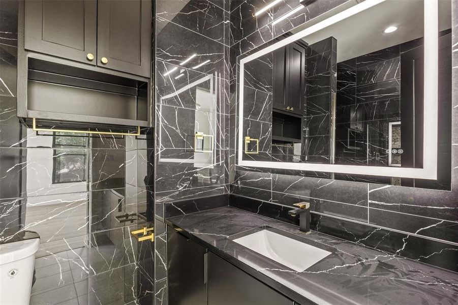 Bathroom featuring black vein-patterned wall and floor tile, a dark-toned vanity with an integrated sink, a large backlit mirror, and a black and gold-toned faucet