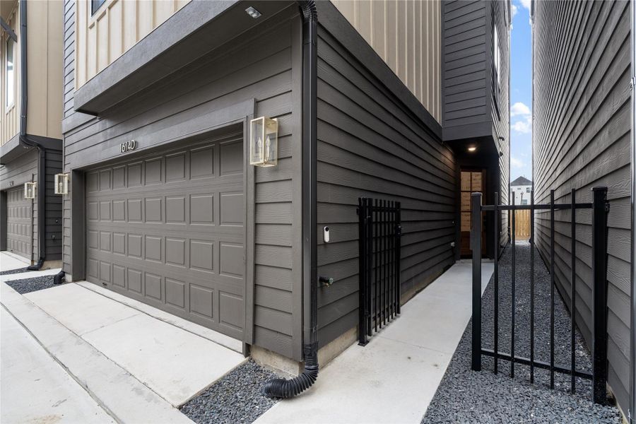 A gated walkway leads to the side entrance, offering both security and privacy. The dark siding and modern light fixtures enhance the sleek design.