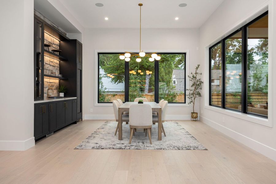 A bright and inviting dining space framed by oversized windows that bring in natural light and views of the outdoors. The neutral palette and clean lines create a calm, modern setting. The highlight is the custom built-in bar with backlit shelving, adding both style and function, making this space perfect for entertaining or relaxed meals at home.