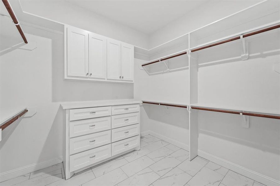 This photo showcases a spacious walk-in closet with built-in white cabinetry and multiple shelves and hanging rods for organized storage. The room features a clean, modern design with light-colored marble-like tile flooring.