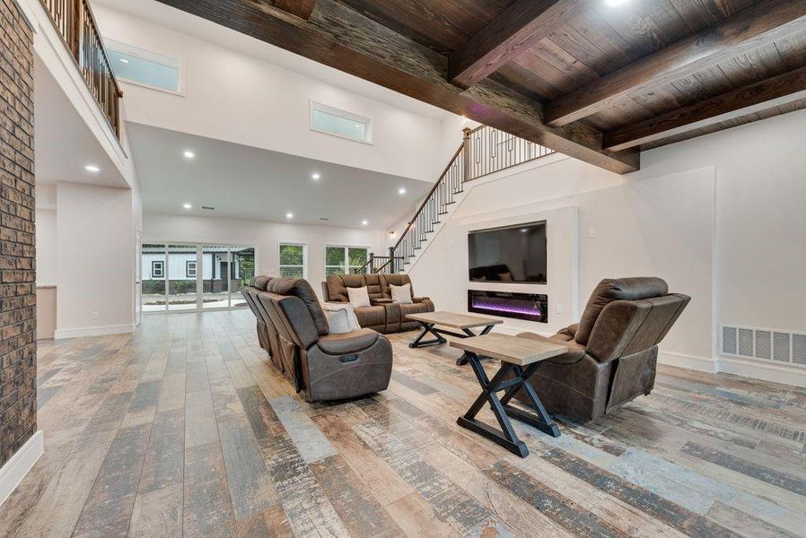 Spacious living area featuring wood-finish flooring, a contemporary fireplace with an integrated television, and a high ceiling with exposed wood beams Spacious living area featuring wood-finish flooring, a contemporary fireplace with an integrated television, and a high ceiling with exposed wood beams