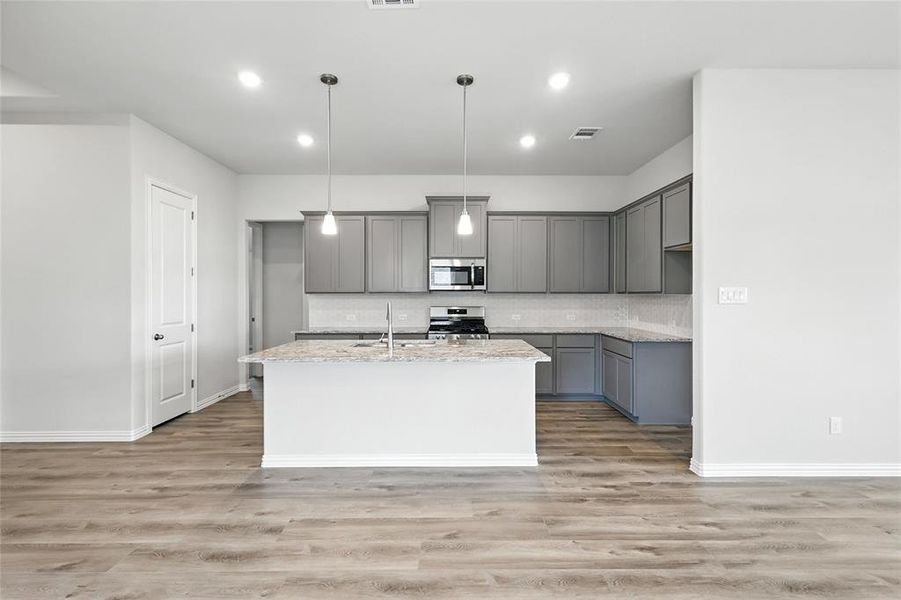Kitchen featuring gray cabinets, light stone counters, hanging light fixtures, an island with sink, and light wood finished floors