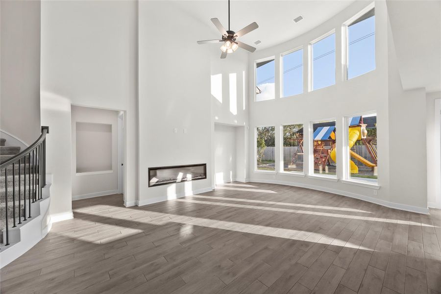 This spacious living area features high ceilings, abundant natural light from large windows, a modern fireplace, and elegant gray flooring. The room flows into a curved staircase with a sleek railing, providing a stylish and open atmosphere. This spacious living area features high ceilings, abundant natural light from large windows, a modern fireplace, and elegant gray flooring. The room flows into a curved staircase with a sleek railing, providing a stylish and open atmosphere.