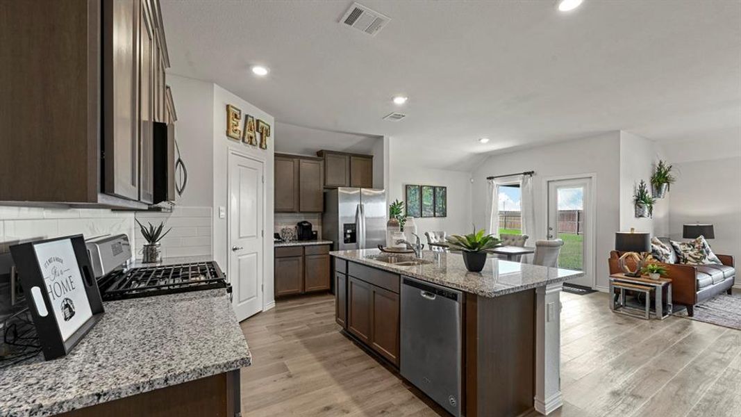 Kitchen with light stone counters, dark brown cabinetry, appliances with stainless steel finishes, open floor plan, and an island with sink