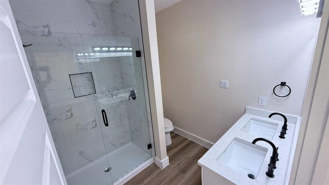 Bathroom featuring a spacious walk-in shower with clear glass enclosure and marble-look tile surround, dual vanity with undermount sinks and black fixtures, light speckled countertop, and wood-finish flooring Bathroom featuring a spacious walk-in shower with clear glass enclosure and marble-look tile surround, dual vanity with undermount sinks and black fixtures, light speckled countertop, and wood-finish flooring