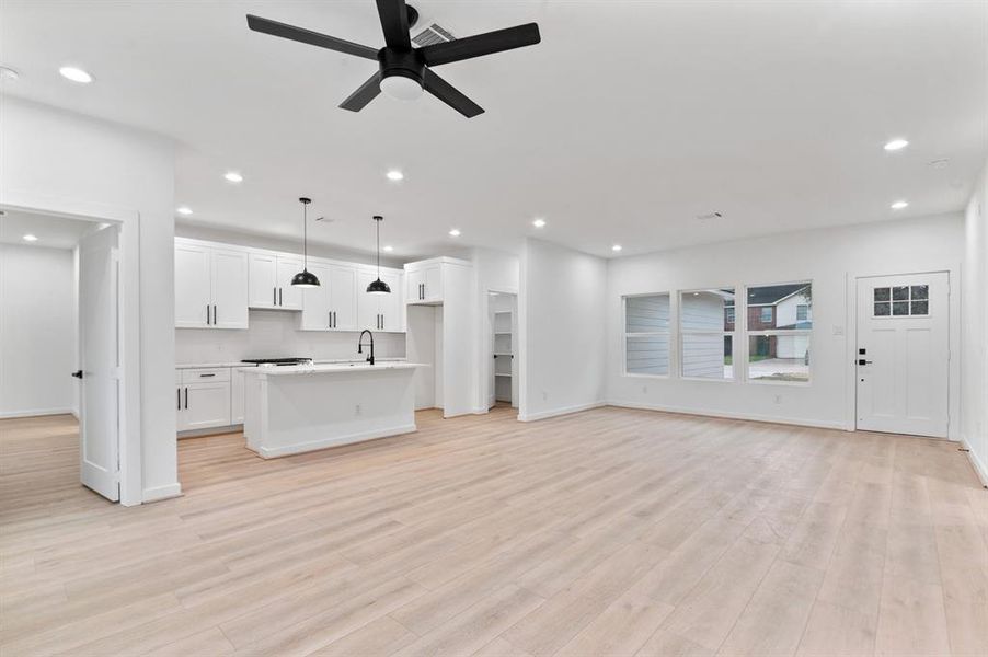 This open-concept living space features a modern kitchen with white cabinets, an island, and pendant lighting. The area is bright with recessed lighting and large windows, complemented by light wood flooring and a ceiling fan. The entry door adds a charming touch.