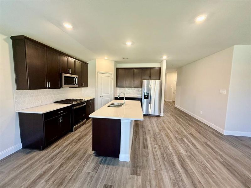 Spacious kitchen featuring dark wood cabinetry, white countertops, stainless steel appliances, a white tile backsplash, and wood-finish flooring Spacious kitchen featuring dark wood cabinetry, white countertops, stainless steel appliances, a white tile backsplash, and wood-finish flooring
