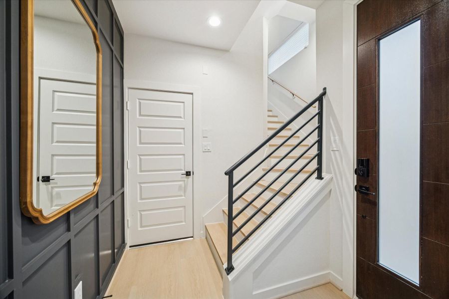 Entryway features a modern aesthetic with LVP light oak style floors, white trim, and a dark accent wall for contrast. The open staircase with black iron railing leads to the upper level, setting a sleek and inviting tone for the home. Entryway features a modern aesthetic with LVP light oak style floors, white trim, and a dark accent wall for contrast. The open staircase with black iron railing leads to the upper level, setting a sleek and inviting tone for the home.