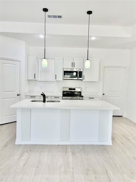 Kitchen with white cabinets, decorative light fixtures, a kitchen island with sink, stainless steel appliances, and light stone countertops