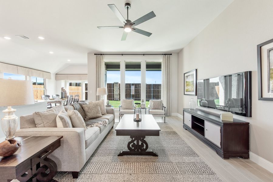 Living room with neutral sofa, ceiling fan, wall-mounted TV, and large windows overlooking backyard Living room with neutral sofa, ceiling fan, wall-mounted TV, and large windows overlooking backyard