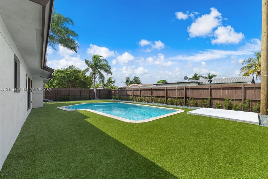 Outdoor living area featuring an in-ground swimming pool, artificial turf, perimeter wood fencing, and a concrete patio slab
