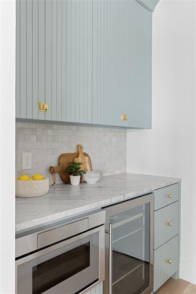 Bar featuring wine cooler, stainless steel microwave, light stone countertops, gray cabinetry, and backsplash