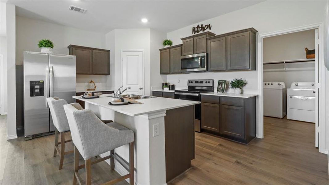 Kitchen with appliances with stainless steel finishes, dark brown cabinetry, washer and clothes dryer, dark wood finished floors, and a center island with sink