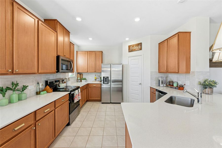 The kitchen features warm wood-finish cabinets, sleek stainless steel appliances, light tile-patterned flooring, a functional peninsula for additional workspace, and a stylish decorative backsplash that adds the perfect finishing touch. Beautiful natural quartz stone countertops enhance the surfaces throughout, adding both durability and refined elegance to the home.