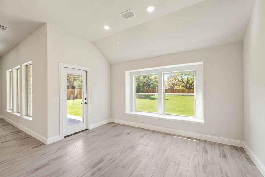 Empty living room with vaulted ceiling, light wood floors, and large windows overlooking a backyard