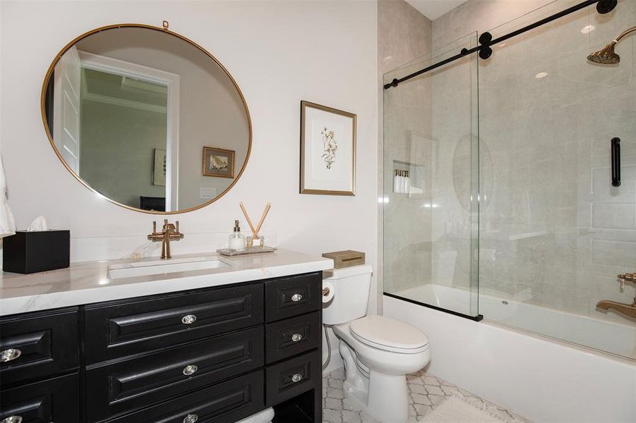 This bathroom features a modern design with a sleek black vanity, marble countertop, and round mirror. The shower has a glass sliding door and elegant brass fixtures, complemented by neutral tile walls and geometric floor tiles.