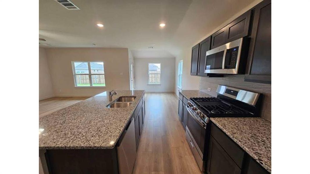 Kitchen featuring an island with a double basin sink, granite countertops, wood-finish flooring, dark cabinetry, and stainless steel appliances including a range and built-in microwave Kitchen featuring an island with a double basin sink, granite countertops, wood-finish flooring, dark cabinetry, and stainless steel appliances including a range and built-in microwave
