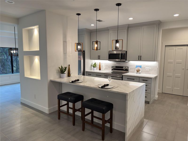 Kitchen with a kitchen bar, stainless steel appliances, gray cabinetry, backsplash, and light stone counters Kitchen with a kitchen bar, stainless steel appliances, gray cabinetry, backsplash, and light stone counters