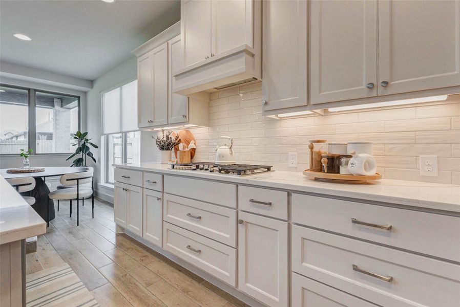 White cabinetry with sleek hardware, a gas stove, and under-cabinet lighting. The light wood flooring complements the bright and airy atmosphere, enhanced by large windows and an adjacent dining nook with stylish seating. White cabinetry with sleek hardware, a gas stove, and under-cabinet lighting. The light wood flooring complements the bright and airy atmosphere, enhanced by large windows and an adjacent dining nook with stylish seating.