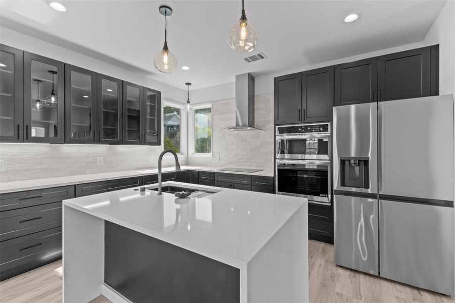 Kitchen with stainless steel appliances, light stone countertops, a center island with sink, decorative light fixtures, and light wood-type flooring