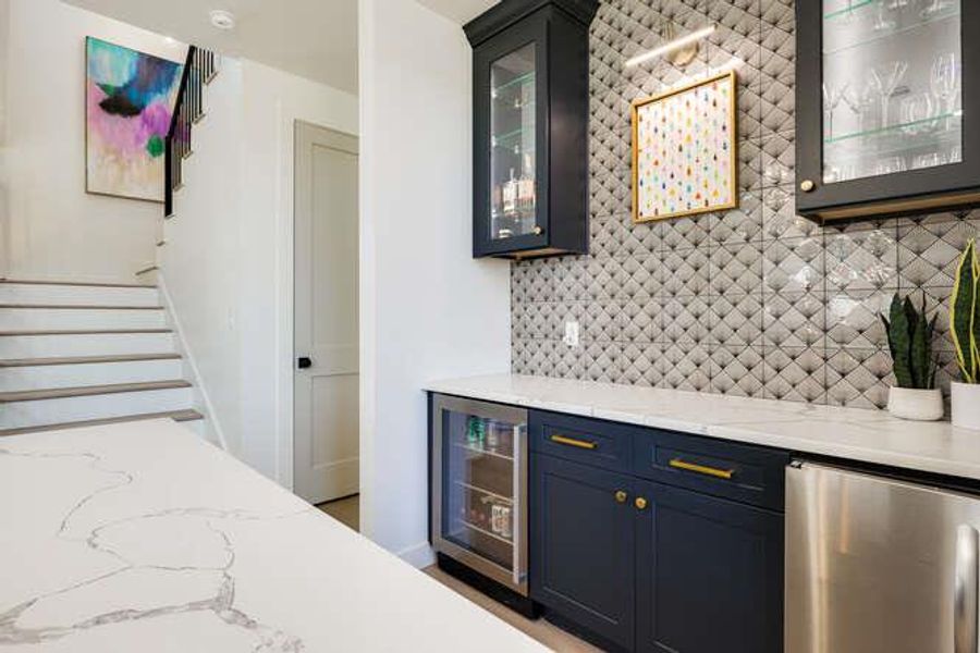 Indoor dry bar featuring decorative backsplash, light stone countertops, stairs, wine cooler, and glass insert cabinets