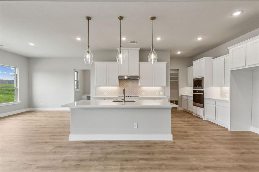 Kitchen featuring stainless steel oven, hanging light fixtures, white cabinetry, and a center island with sink