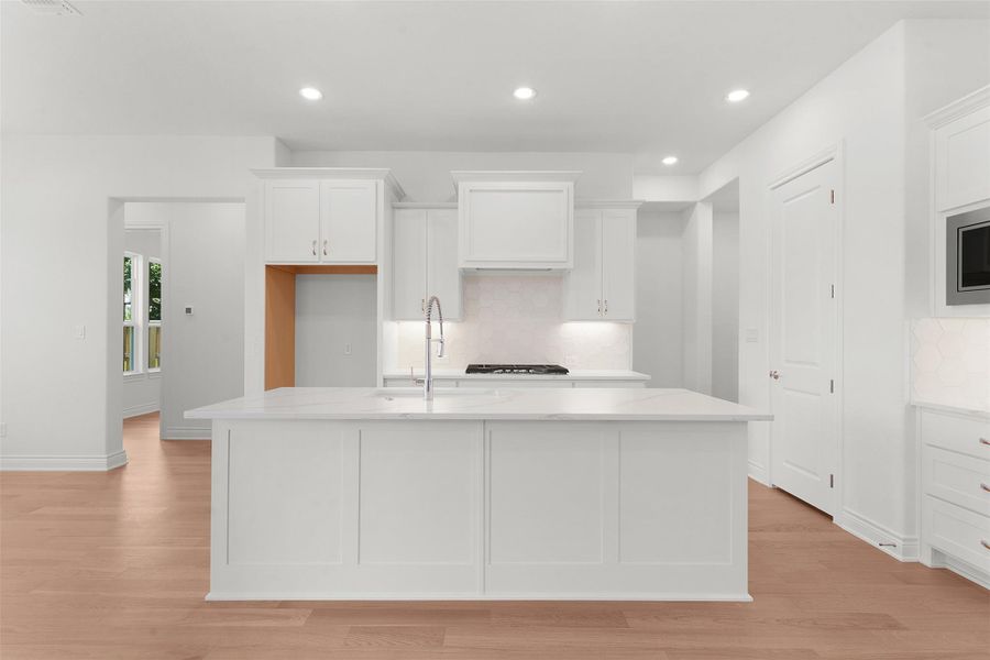 Modern kitchen featuring a central island with an integrated sink, white shaker cabinetry, quartz countertops, a tile backsplash, and light wood-finish flooring
